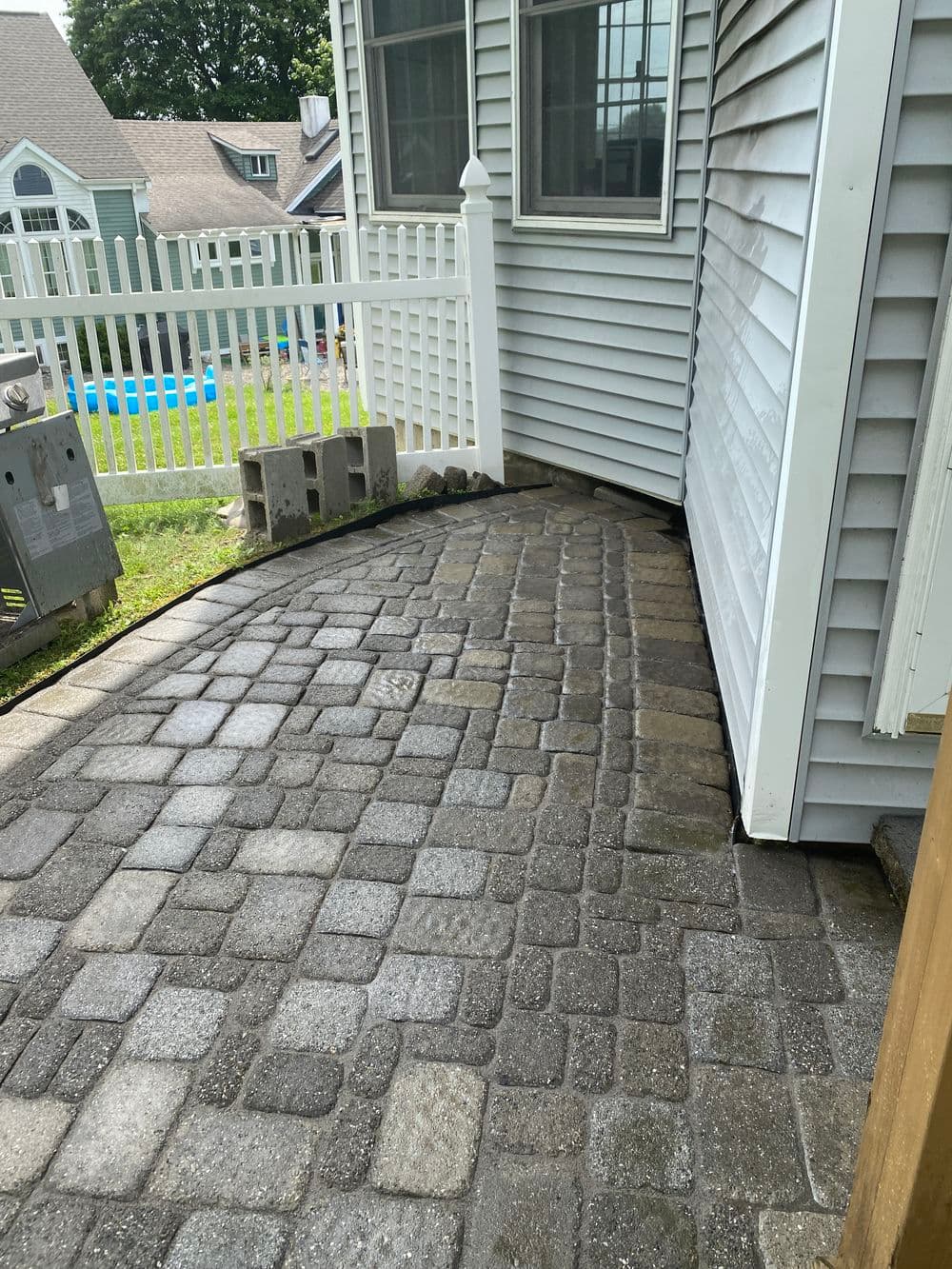 Paved patio area beside a house with a white fence and green lawn in the background.